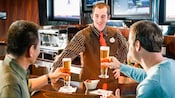 A smiling Cast Member serves 2 tall glasses of beer to 2 men at a bar