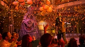 Famílias assistem a um show de Halloween com uma árvore assustadora, lanternas de abóbora, luzes laranjas e confete Famílias assistem a um show de Halloween com uma árvore assustadora, lanternas de abóbora, luzes laranjas e confete