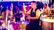 Un barman maniobra con una botella en el aire mientras prepara un coctel detrás de una barra