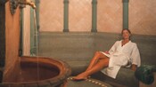 A woman in robe relaxes in the steam of a tiled hammam that contains a running fountain