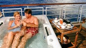 A couple sit in a private hot tub together, clinking champaign glasses out on an open deck overlooking the sea