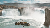 Two waterfalls with a large stone formation between them and rolling hills without in the background