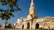 Una pared de piedra con tres puertas separa una plaza de una ciudad con edificios modernos y coloniales