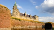 A castle with spires and raised ceilings sits above grass and a brick wall lining a moat