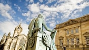 A weathered statue of a man holding a book in front of old buildings