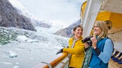 A woman holding binoculars smiles with a young girl on a ship deck in a mountainous setting with floating ice caps