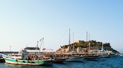 Six fishing boats bearing the Turkish flag sit in a row in front of a round island circled by stone walls