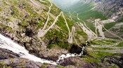 A narrow strip of of road zig-zags down a mountain side and over a gorge cut by a flowing waterfall