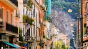 Buildings with balconies holding flowers line a street with a cliff face in the background