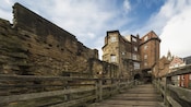 A wooden bridge leading past the ruins of a brick wall to a large building