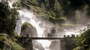 Three people stand on a bridge looking out with a waterfall cascades down rocks behind them
