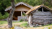 A building with a raised roof sits behind a smaller structure built of interlocking logs