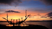 Silhouetted sculpture in the shape of a boat sits on a platform at sunset