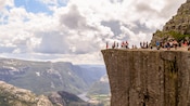 An assortment of people sitting and standing above a sheer cliff overlooking a valley