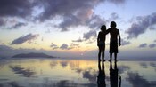 A boy puts his arm around a girl, as they walk in silhouette on the shoreline at sunset