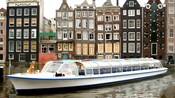 A glass covered canal boat glides through the water in front of canal houses with gabled facades