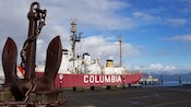 A large anchor on a dock near a boat with a sign on it that reads Columbia