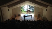 A piano sits on a stage lit by natural light while an audience anticipates a performance