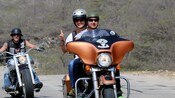 A couple rides a Harley Davidson motorcycle while another biker follows behind them