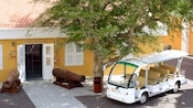 2 rusty cannons, a shuttle bus and a tree in front of a building