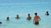 A group of vacationers relax with drinks in hand and they stand in shallow, clear ocean water