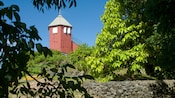 A tower rises out of thick trees on a hill with a stone wall