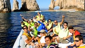 A group of people in an inflatable raft smile and wave to the camera with Los Arcos in the background