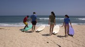 Three young people standing on the beach watch as an instructor demonstrates how to stand on a board