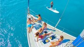 2 men and 3 women sunning on a boat deck while a 3rd man tries standup paddleboarding in the sea