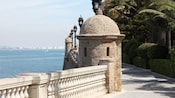 A fishing pole on a bridge lined with gas lamps and domed brick fixtures, looking out toward the sea