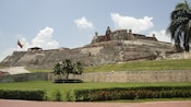 A flag waves outside an ancient stone fortress on a hill, with a few palm trees nearby