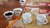 Pequeñas tazas de café molido y granos de café colocadas en una mesa con una taza, un lápiz y manteles individuales