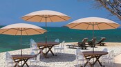 An ocean-side deck with 3 tables and chairs, umbrellas and chaise lounges