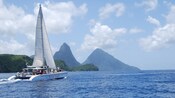 Un catamarán con una vela alta que lleva a los pasajeros por el océano hacia las montañas puntiagudas