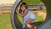 A young girl peeks out from the inside of a giant tire in a grassy farm area
