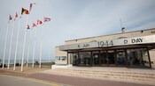Bandeiras próximas a uma construção com uma placa que diz 6 Juin 1944 D Day