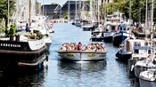 A boat with passengers steers through a waterway flanked by other sea vessels