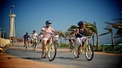 A group of bicycle riders smiling as they pass a lighthouse next to ocean.