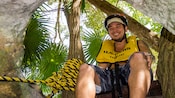 A young man wearing a life vest smiles while sitting in a tree