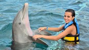 A girl in waist high water stands parallel to a dolphin while holding on to its pectoral fins