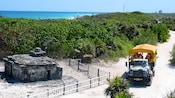 A truck with a canopy over its bed carries passengers past the ruins of an old structure with a low fence