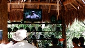 In a thatched roof hut, people watch a television screen with video of a man in safari clothes