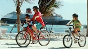 Two children and a woman ride bikes across the beach with a Disney Cruise Line ship in the background