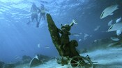 Snorkelers encounter Mickey Mouse underwater straddling a piece of wood next to a ship steering wheel