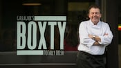 A man smiling next to a window sign that says gallaghers boxty totally Irish