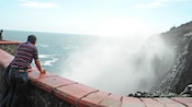 A man looks over a railing at an erupting geyser that is spraying from the sea