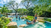 A family stands in a cascading pool by trees, buildings, a river and rocks