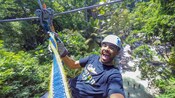 A man hangs from a zip line above a jungle and people standing in a body of water