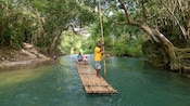 A guide propels a bamboo raft through a forested river while the two women passengers enjoy the ride