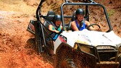 Uma mulher e uma criança usando óculos de proteção e capacetes sorriem enquanto dirigem por um terreno muito lamacento em um buggy